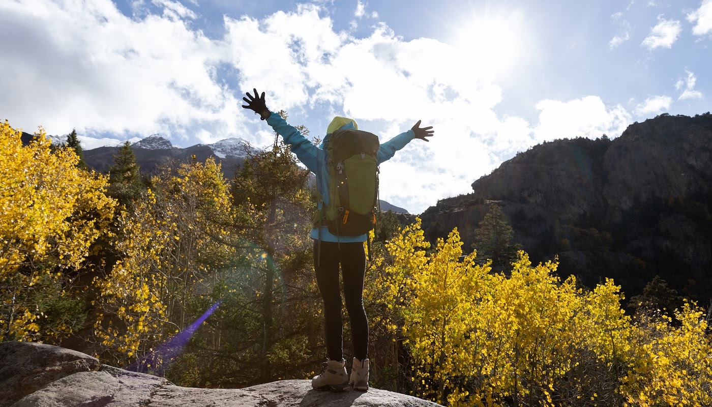 Image: backpacking hiker on top of cliff overlooking trees, blue sky, and mountain range