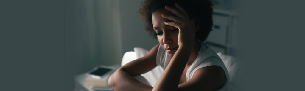 Image of woman sitting in her bed visibly under stress with her hand on her brow