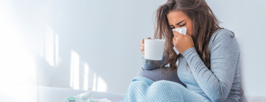 Image of woman with mug in her hand, sneezing, as she sufferers through cold & flu season 