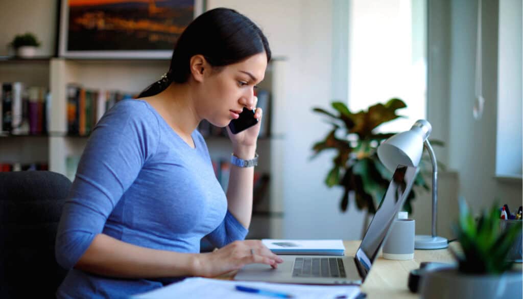 A woman sits at a desk, talking on the phone while looking at her laptop. With a lamp and plants in the background, she projects an inviting "contact us" vibe as she navigates her tasks with ease.