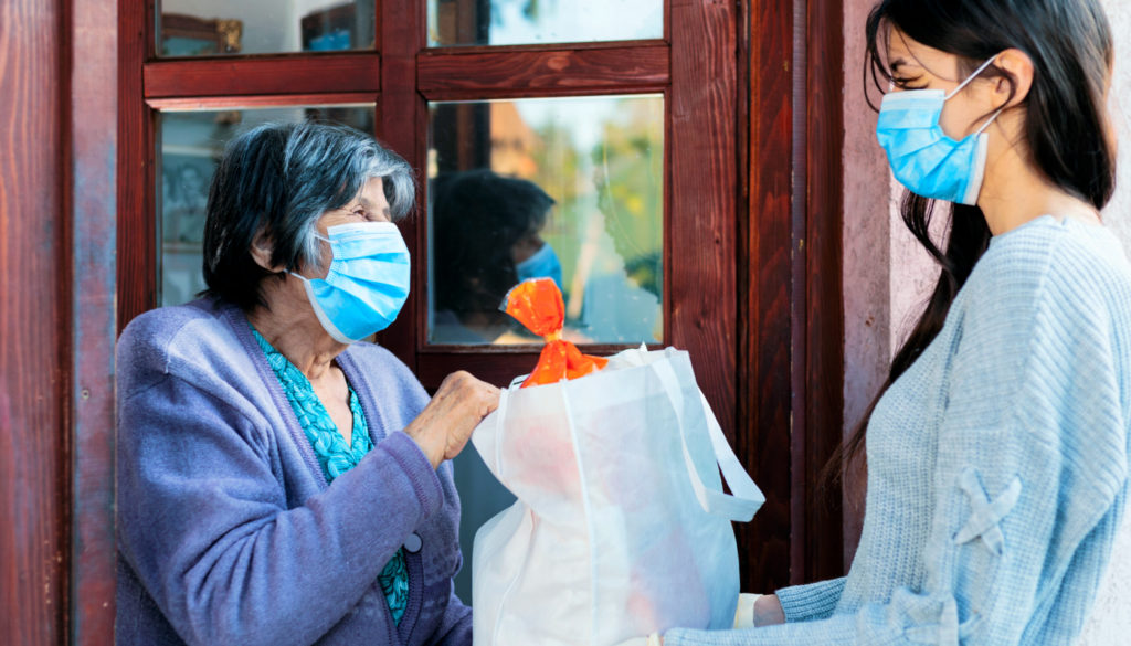 Image of a woman with mask giving a gift to an older woman in front of her home