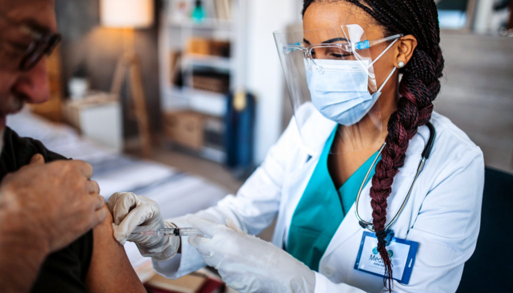 Image of woman nurse in ppp gear administering a vaccination to a male patient