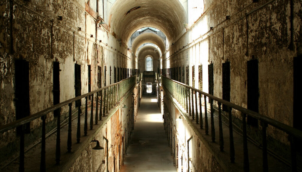 Long, empty prison corridor with high arched ceiling, rusted metal railings, and worn walls; sunlight streams through windows at the far end, casting eerie shadows perfect for a Halloween-inspired travel nursing contract photo.