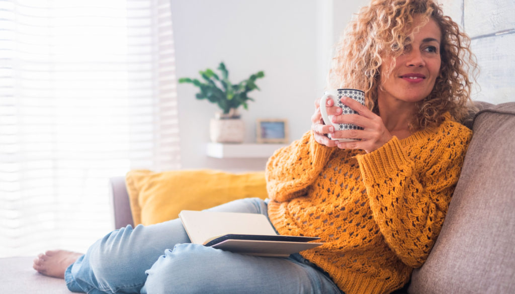 Image of young woman smiling on her couch while nursing a mug of her favorite drink