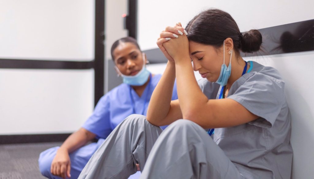 image of nurse sitting with head resting on hands while another nurse consoles her