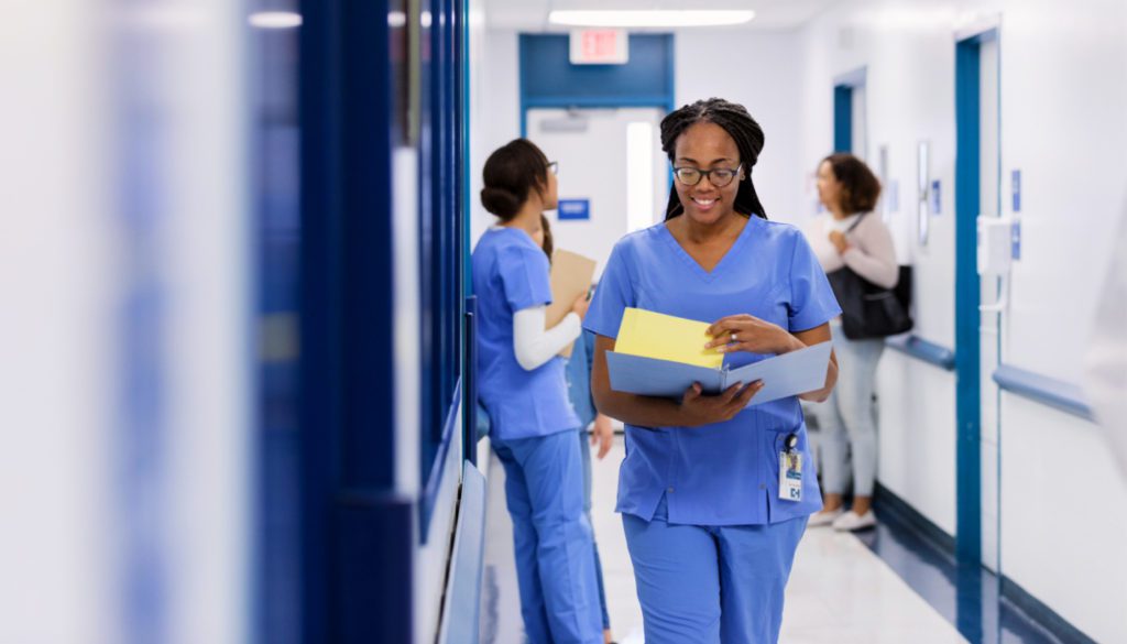 This is an image of a registered nurse walking towards the foreground in scrubs, smiling while flipping through a patient's chart.