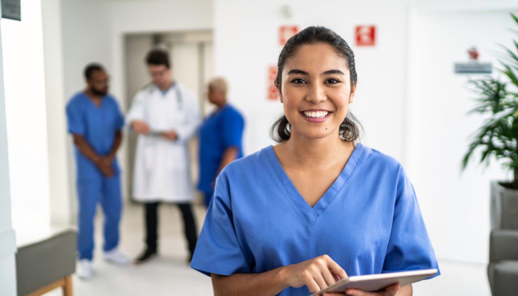 photo of a nurse smiling at the camera with three healthcare professionals talking in the blurred background