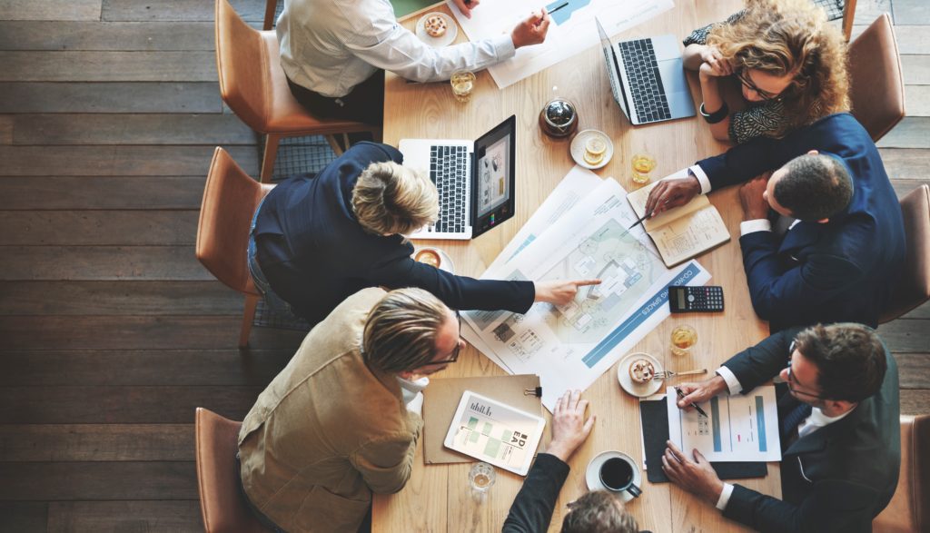 overhead image of professionals at a conference table filled with laptops, notepads and charts, discussing business