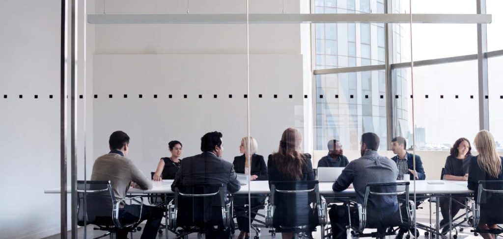 A group of people in business attire is seated around a conference table in a modern office with glass walls, discussing innovative workforce solutions.