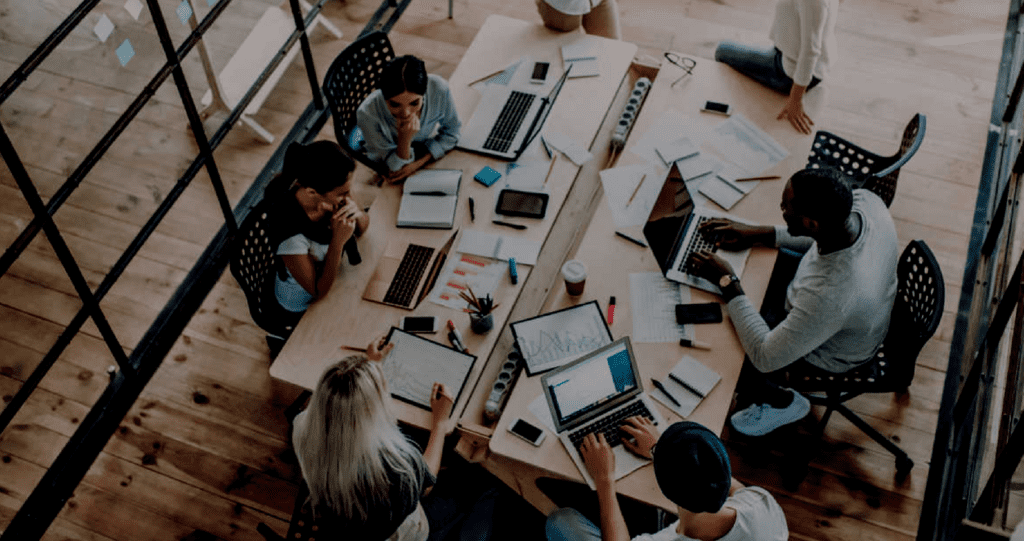overhead image of professionals at a conference table filled with laptops, notepads and charts, discussing business