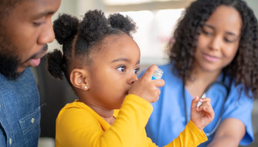 A candid photo of a toddler using her inhaler under the supervision of her father and a nurse