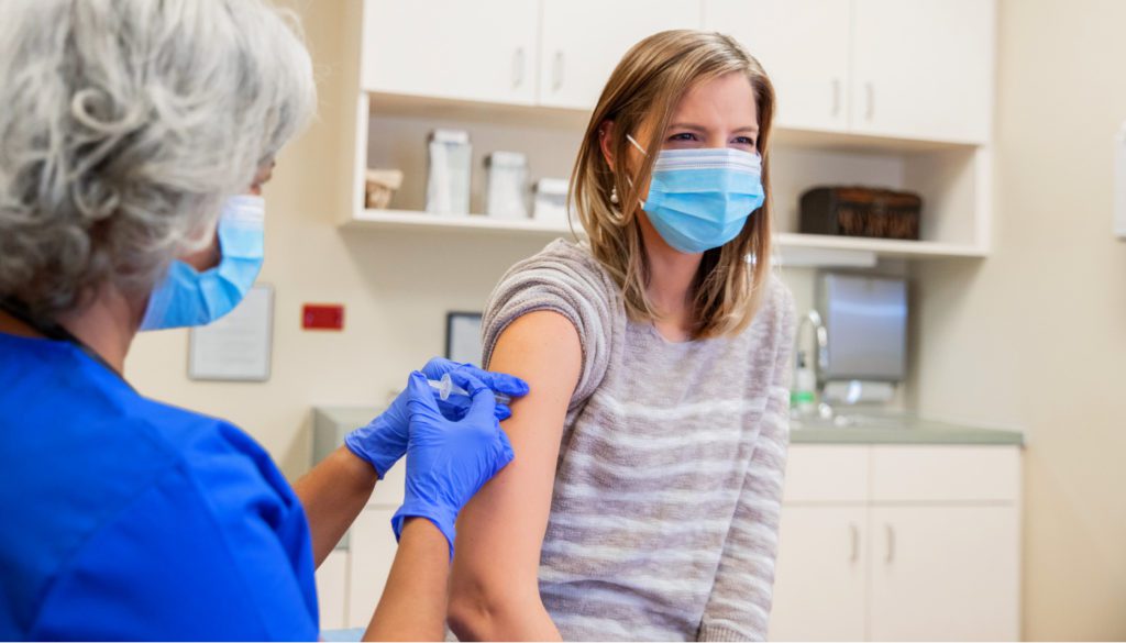 image of a woman nurse in a mask and scrubs administering a needle to a woman patient wearing a mask.