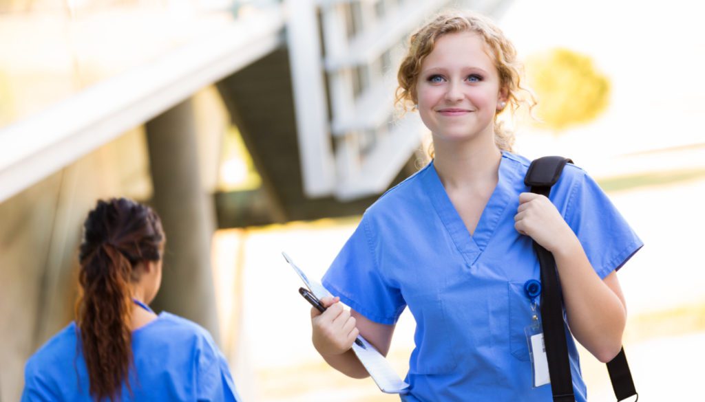 Image of a nursing student smiling, wearing blue scrubs and carrying a bag over one shoulder while holding a pen and papers in her right hand.