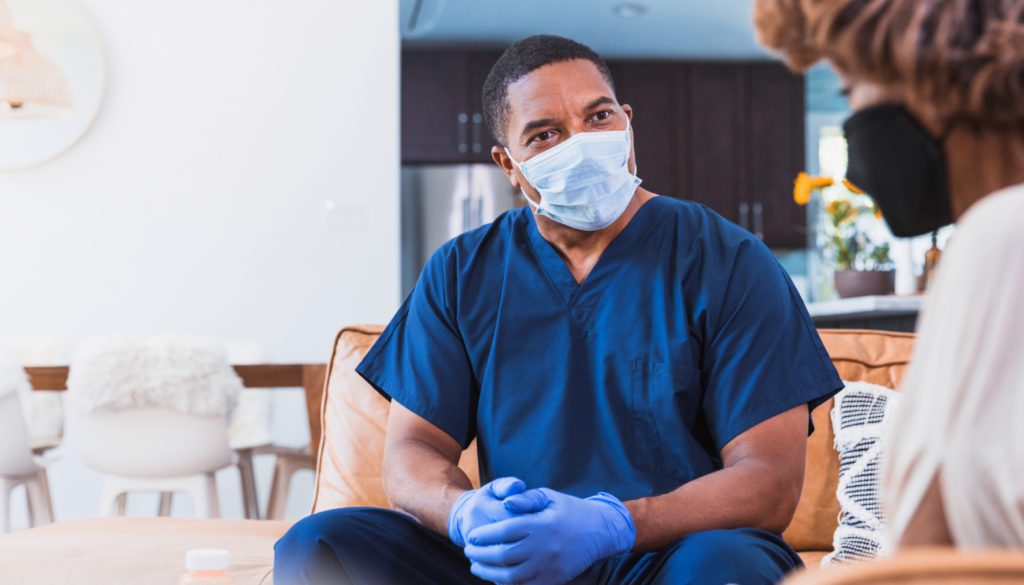 image: medical professional wearing a mask and gloves as he speaks with a woman in her home