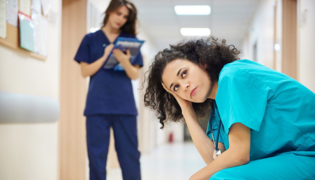 A dedicated medical professional in turquoise scrubs sits in a hallway, resting their head on their hand, as they navigate the demanding world of healthcare. Meanwhile, a colleague in blue scrubs stands nearby with a clipboard, ready to discuss innovative workforce solutions.