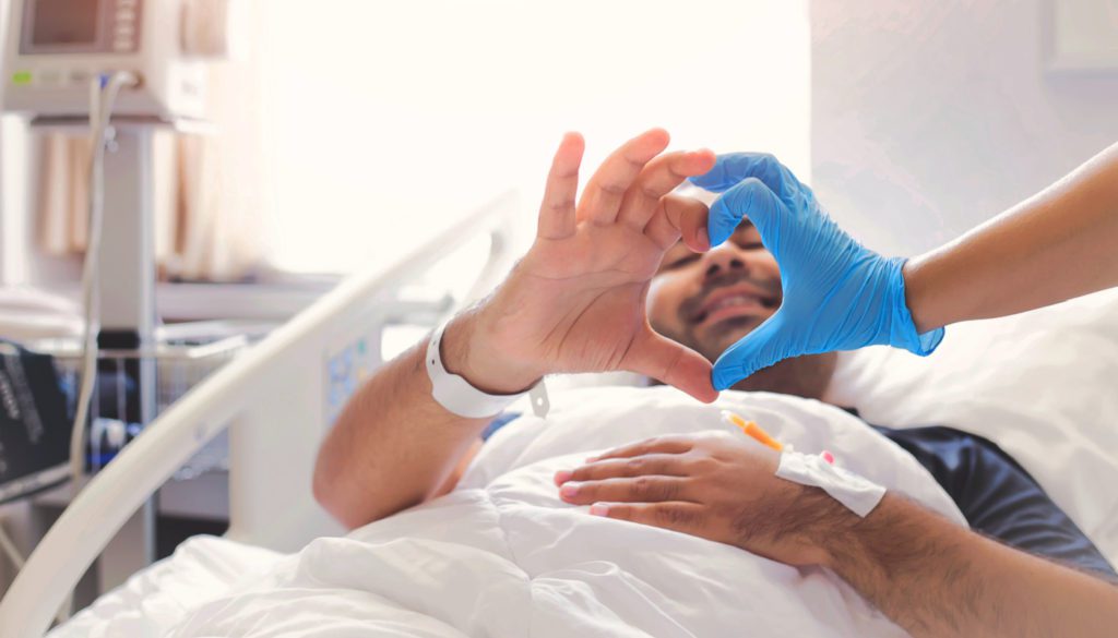 image of a patient in a hospital bed gesturing half a heart with one hand while a gloved hand from a nurse completes the gesture