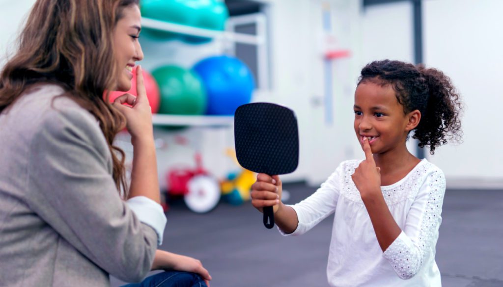 image: woman smiling as she teachers a young girl holding a mirror to smile as well