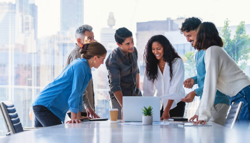 Image: Team of business people working together in a board room. They are all standing and talking. There is technology on the table including a laptop computer. Ethnically diverse group. There is also some paperwork.