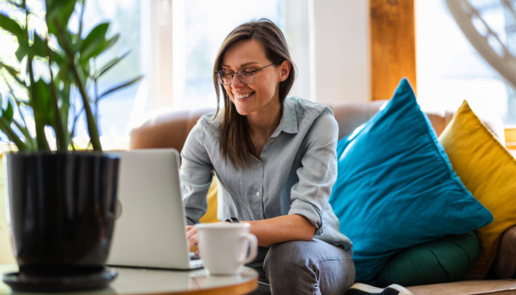 Banner: Woman smiling while sitting on a couch working on a laptop, depicting travel SLP Audiology