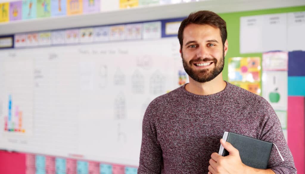 A man with a beard stands in a colorful classroom, smiling and holding a closed book, like a dedicated school teacher, with charts and drawings visible on the blurred whiteboard behind him. School professional smiling because there are summer jobs for school teachers, paraprofessionals, and school nurses across the country.