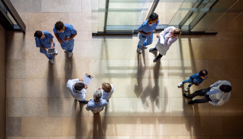 Aerial view of healthcare professionals in scrubs and lab coats standing and talking in a hospital corridor with tiled floors, illustrating the collaborative spirit of workforce solutions.