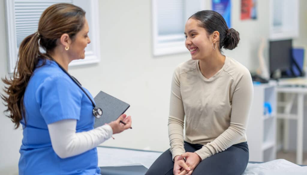 A Native American nurse talking to a healthcare patient on a bed.