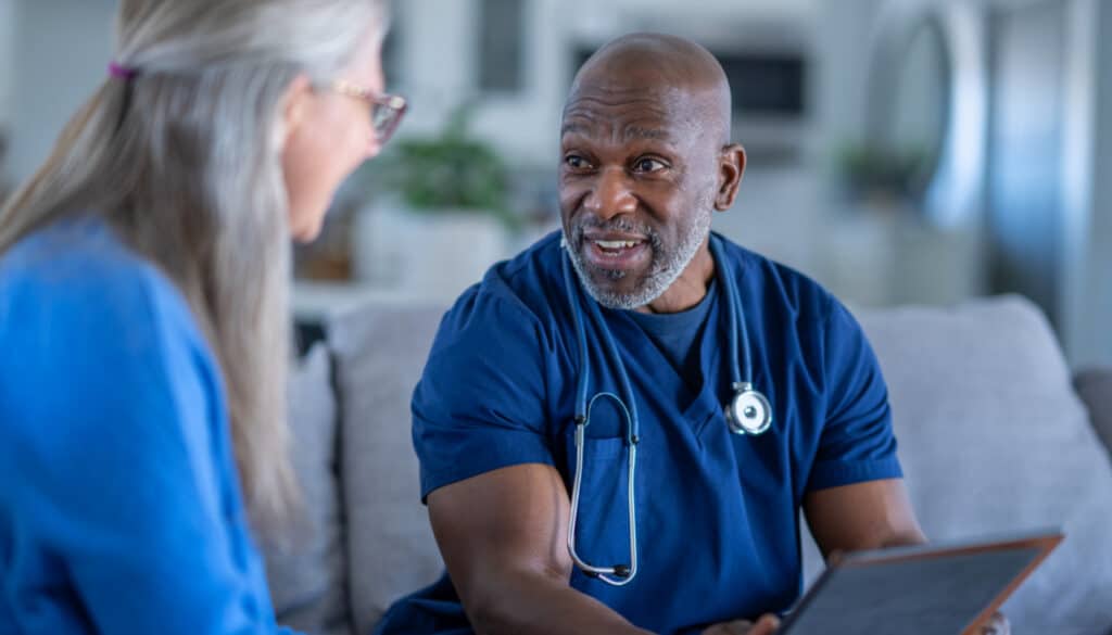 A home health nurse talking to a patient on a couch in their home.