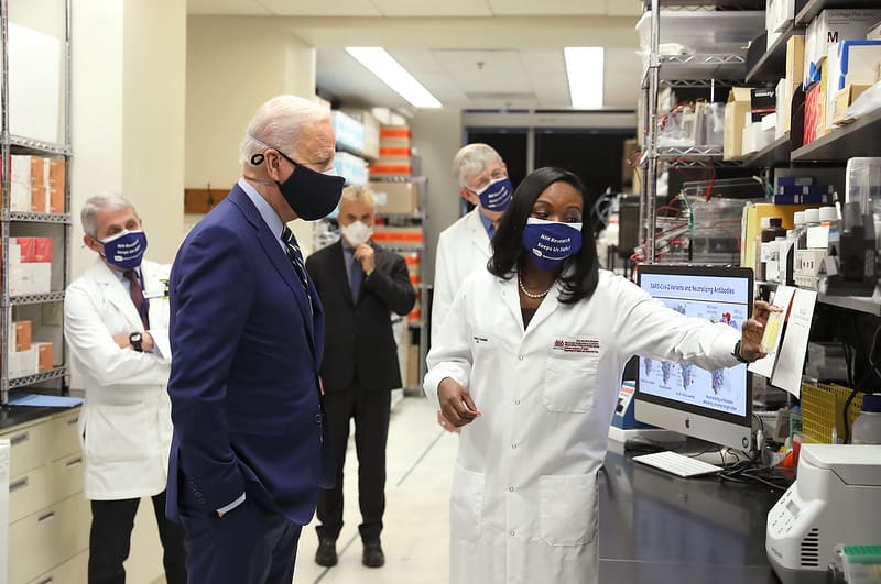 President joe biden inspects a laboratory with people wearing face masks.