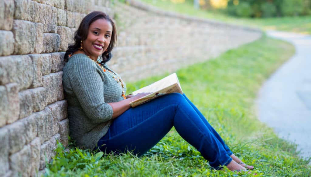 A young black woman reading a book on the side of a stone wall.