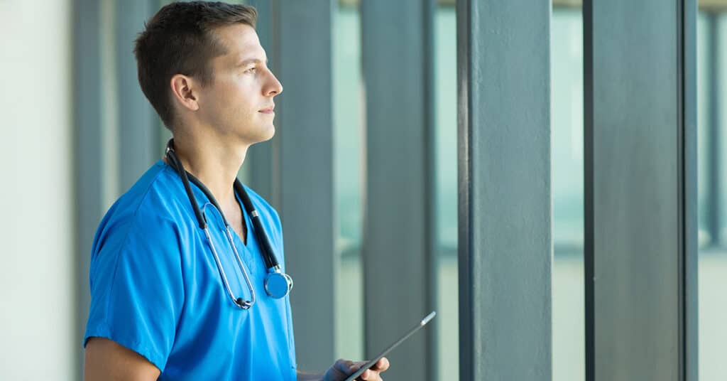A male nurse standing in a hallway holding a clipboard.