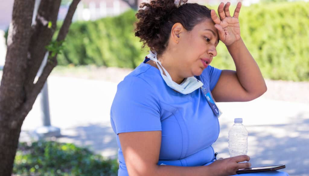 A tired nurse in blue scrubs sits outside, resting her head on her hand, with a face mask around her neck and holding a water bottle.