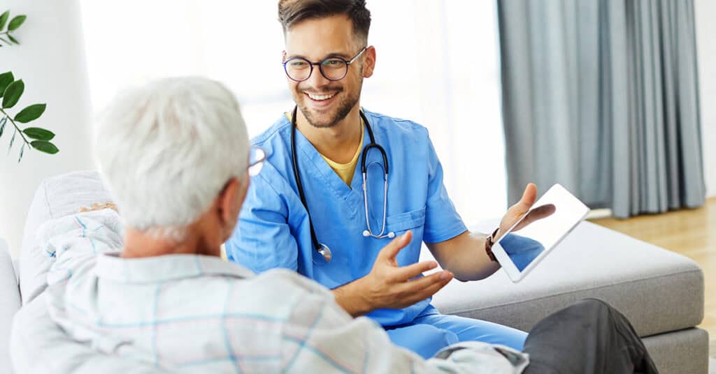 A male nurse in scrubs discussing with an elderly male patient on a sofa, holding a digital tablet in a bright, modern living room.