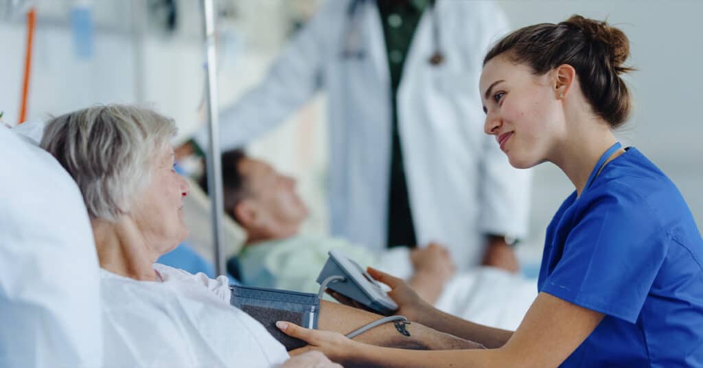 A nurse attentively listens to an elderly patient while holding a tablet in a hospital ward.