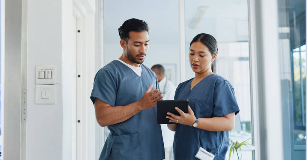Two healthcare professionals in scrubs discussing information on a tablet in a hospital corridor.