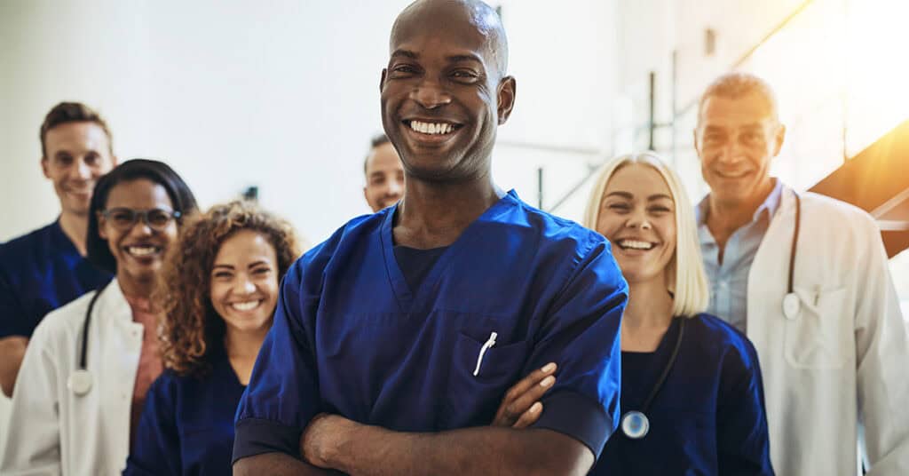 A group of diverse healthcare professionals smiling in a hospital setting, with a black male nurse in blue scrubs at the forefront.