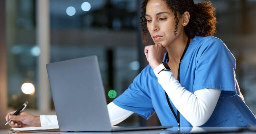 A healthcare professional in scrubs using a laptop and taking notes at a desk in a modern office setting.