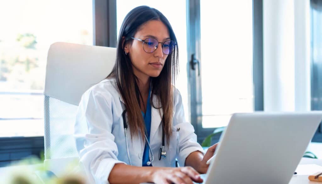 A female doctor in a white coat and glasses using a laptop in a bright office setting.