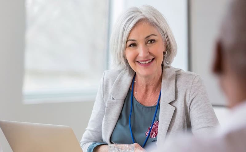 A smiling senior woman with gray hair in a business meeting, wearing a gray blazer and blue top, discussing with a colleague.