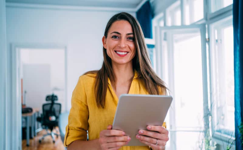 A woman in a yellow shirt smiles, holding a tablet in a bright room with blue curtains, seamlessly engaging in direct sourcing.