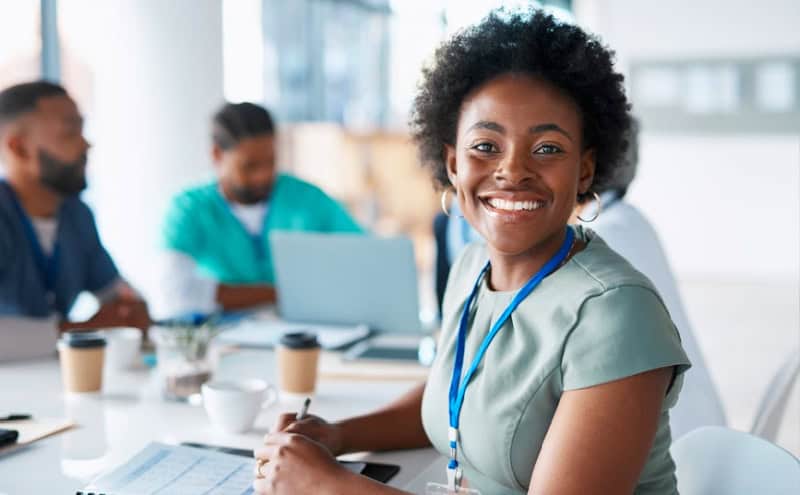 Smiling black woman with colleagues in a business meeting. she wears a lanyard and sits at a table with laptops and coffee cups visible.