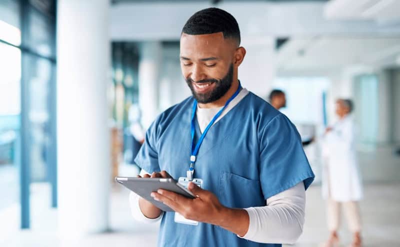A male nurse in blue scrubs using a tablet with a smile, standing in a bright hospital corridor.