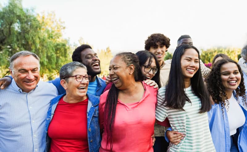 A diverse group of people are standing outdoors, arm in arm, smiling and laughing together. Trees and greenery are visible in the background.