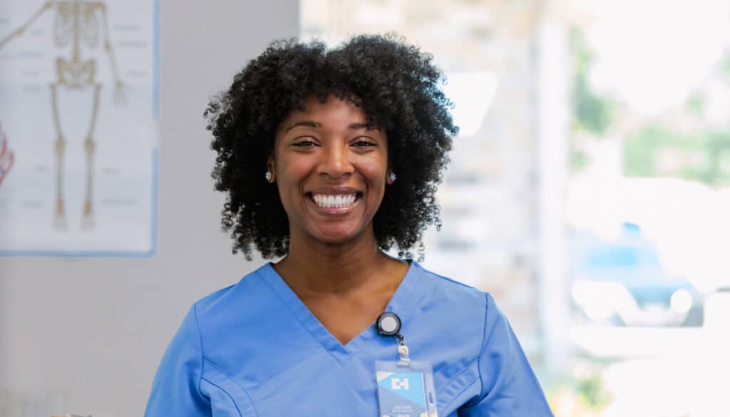 A smiling black female nurse with curly hair, wearing a blue scrub and a stethoscope around her neck, standing in a clinic.