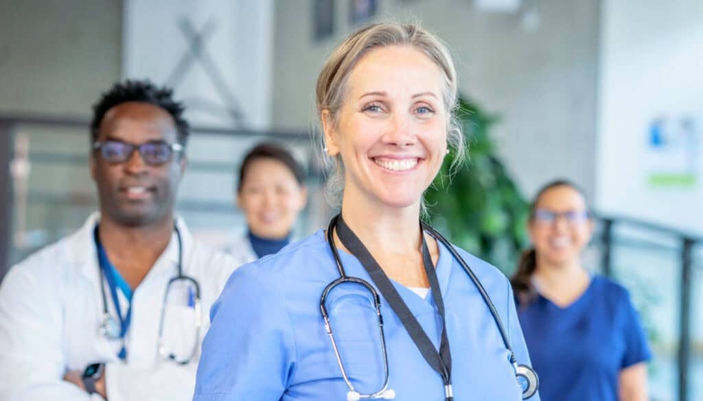 A smiling APRN nurse in a blue scrub top stands in the foreground with three other nursing staff members in the background, exemplifying the dedication typical of those in travel nursing.