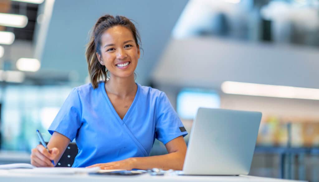 A nurse wearing blue scrubs sits at a desk with an open laptop, writing on paper and smiling at the camera.