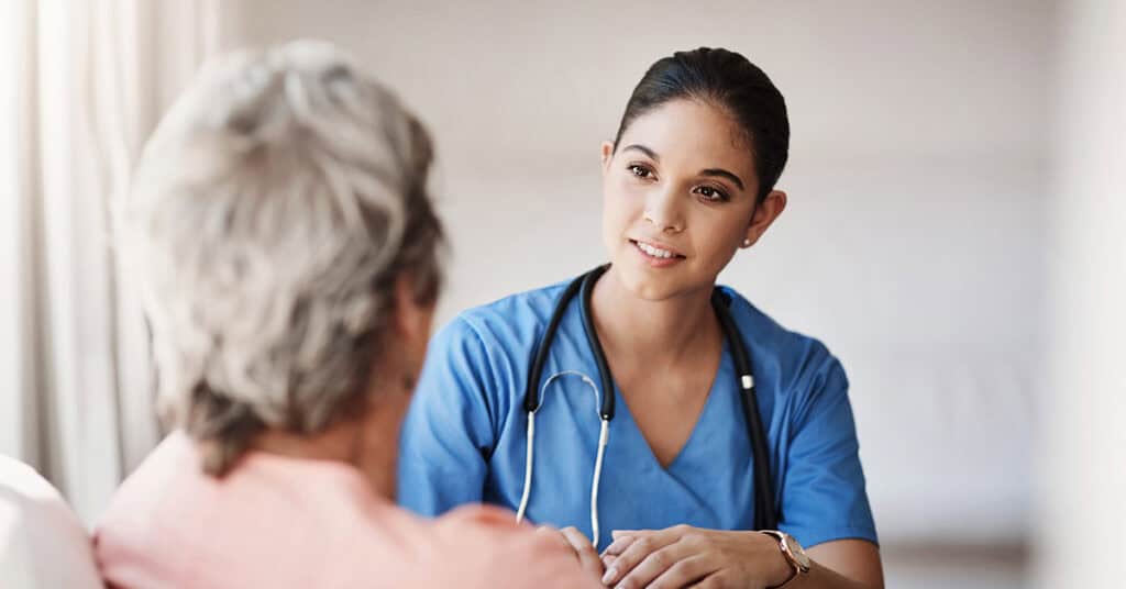 A female healthcare worker in blue scrubs, with a stethoscope around her neck, listens attentively to an elderly person in a pink shirt. They are engaged in a conversation in a well-lit room.