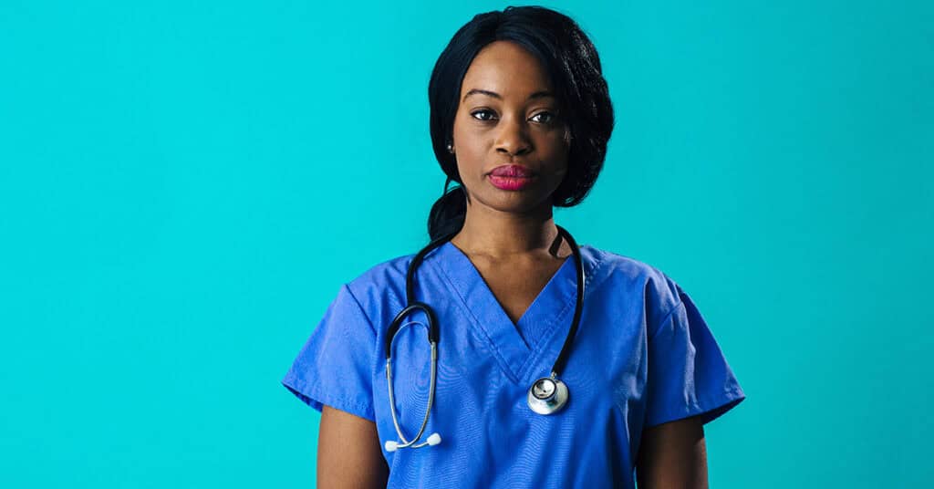 A woman wearing blue scrubs and a stethoscope stands against a turquoise background.