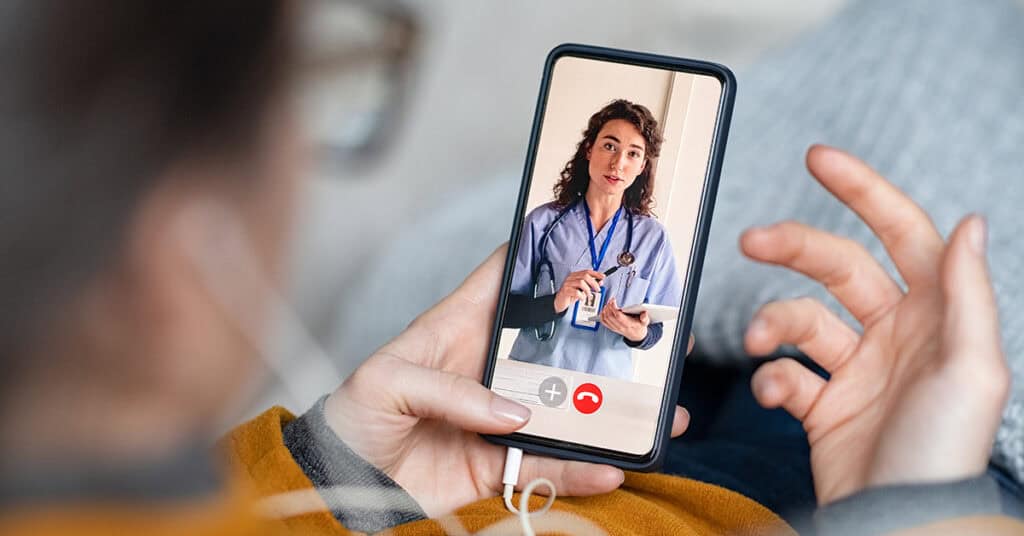 Person in a yellow sweater holding a smartphone showing a video call with a female doctor wearing a stethoscope and blue scrubs.