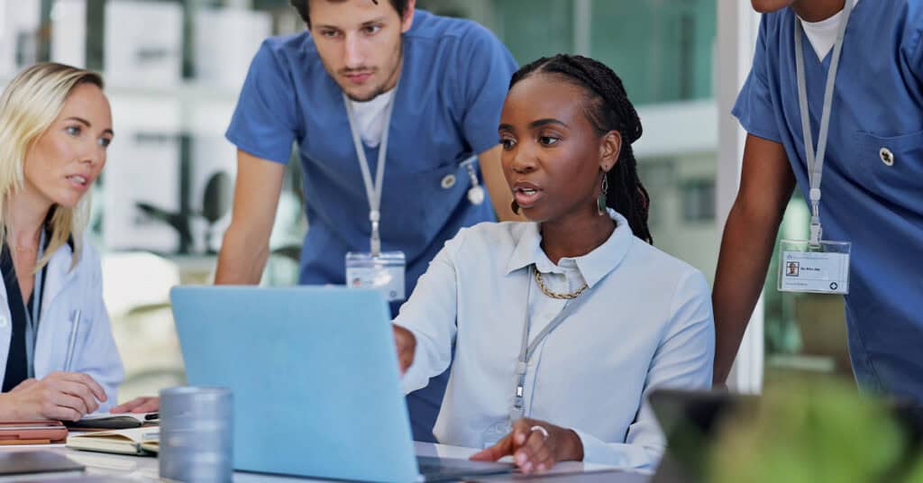 A group of healthcare professionals in uniforms is gathered around a table, engaging in a discussion while looking at a laptop. One woman is pointing at the screen, leading the conversation.