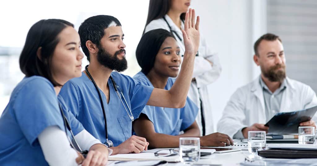 Healthcare professionals in scrubs sit at a conference table with documents, water, and a clipboard, while one person raises their hand, and another stands in the background.